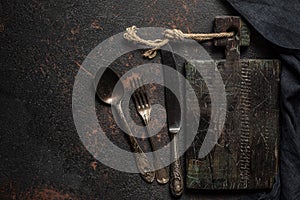 Old wooden cutting board, vintage cutlery and towel on black stone kitchen table. Top view flat lay with copy space