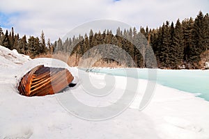 Old wooden destroyed flooded fishing boat lie in snow on shore of ice-covered frozen river in winter, snow in cold morning at