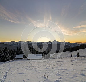 Old wooden house, hut and barn in deep snow on mountain valley, spruce forest, woody hills on clear blue sky at sunrise copy space