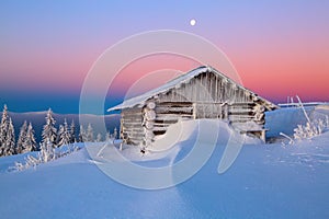 Old wooden hut. Huge snowdrifts around. Background of the high mountains. High fairtrees freezed with snowflakes.