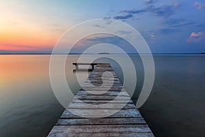 Old wooden jetty, pier reveals views of the beautiful lake, blue sky with cloud. Sunrise enlightens the horizon with orange warm