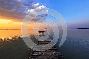 Old wooden jetty, pier reveals views of the beautiful lake, blue sky with cloud. Sunrise enlightens the horizon with orange warm