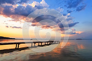 Old wooden jetty, pier reveals views of the beautiful lake, blue sky with cloud. Sunrise enlightens the horizon with orange warm