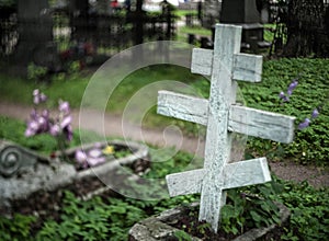 Old wooden orthodox grave cross in a cemetery with a blurry background with flowers and grave fences