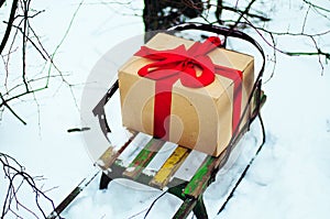 Old wooden sleigh with a gift in golden paper box wrapped red gift ribbon, are in the winter forest, snow, trees near. Wooden sled