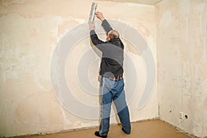 The old worker plastered the wall in the apartment. A man in dirty clothes with a large spatula performs the puttying of the wall