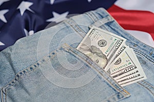 Old worn jeans are lying on flag United States America and there are hundred dollar bills in pocket