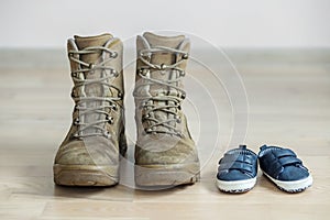 Old worn military boots and baby shoes on wooden floor. Concept of military father and family