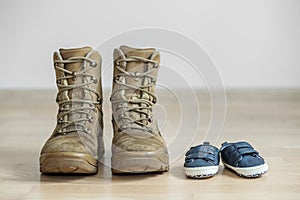 Old worn military boots and baby shoes on wooden floor. Concept of military father and family