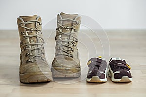 Old worn military boots and children`s shoes on wooden floor. Concept of military father and family
