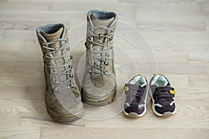 Old worn military boots and children`s shoes on wooden floor. Concept of military father and family