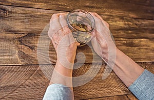 Old wrinkled hands holding jar with coins, wooden background.