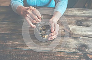 Old wrinkled hands holding jar with coins, wooden background.