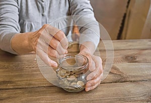 Old wrinkled hands holding jar with coins, wooden background.