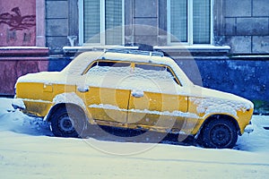 Old yellow car covered with snow parked on a snowy sloping street in winter city