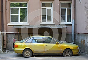 An old yellow car with deflated wheels is parked against the wall of a residential building photo