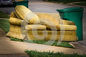 Old yellow couch with cushions sits on a curb in residential area next to a recycling cart waiting for bulk waste hauler pickup on