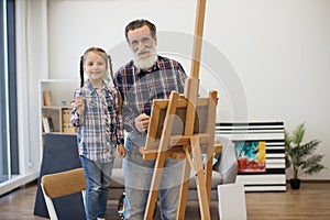 Older adult with girl posing behind easel with cloth in room