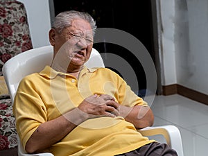 Older Asian men sitting on a chair at living room with heart attacks.Both old man`s hands on breast because of hard breathing