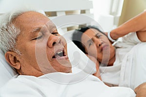 Older Asian woman laying open her eyes and Cover ears with her hands in bed beside her husband who snores and Makes noise, Marital