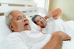 Older Asian woman laying open her eyes and Cover ears with her hands in bed beside her husband who snores and Makes noise, Marital