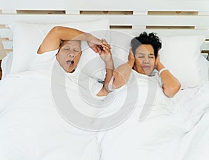 Older Asian woman laying open her eyes and Cover ears with her hands in bed beside her husband who snores and Makes noise, Marital