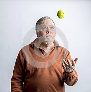 Older bearded man in orange sweater tossing green tennis ball