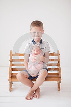 The older brother holds in his hands a newborn sister in a pink cocoon and sits on a wooden bench, brother and sister