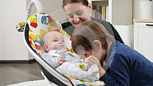Older brother and smiling mother playing with little baby boy rocking in electric rocking seat. Child development and