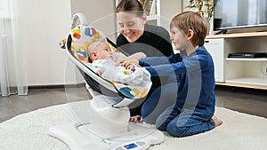 Older brother and smiling mother playing with little baby boy rocking in electric rocking seat. Child development and