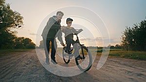 The older brother teaches the younger to ride a two-wheeled bicycle at sunset.