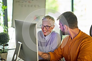 An older business woman and a young male colleague talking about a computer screen content they watching together at workplace.