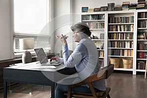 Older businesswoman sit at desk use smartphone talks on speakerphone