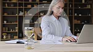 Older businesswoman working on laptop, focus on hourglass