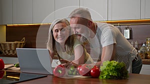 Older Caucasian couple middle-aged spouses family using laptop device at home kitchen choosing food delivery fresh