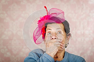 Older cute hispanic woman wearing blue sweater, large pink ribbon on head loking into camera covering her mouth with one