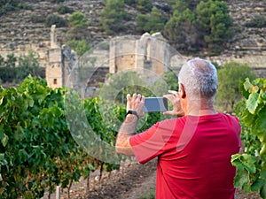 Older farmer in a vineyard with a ruined monastery in the background, taking a picture with his mobile phone