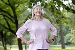 An older gray-haired woman stands in a park in a supportive outfit, holds her hands on the side of her body and closes