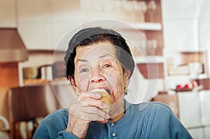 Older hispanic happy woman wearing blue sweater sitting in front of camera having a bite off cupcake