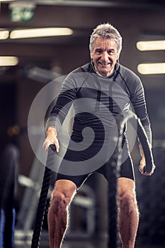 Older man with headphones exercises with ropes in a fitness training center