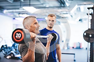 Older man lifting weights, supervised by gym assistant.