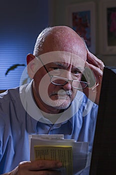 Older man looks worried as he pays bills online, vertical