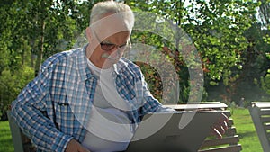 An older man sitting on the bench with a laptop in the garden.