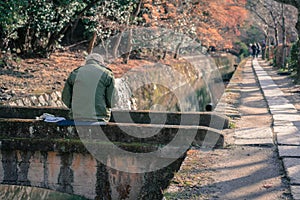 An older man sitting on a stone bridge across a water canal painting picture in the Philosophers walk in Kyoto, Japan. Autumn