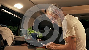 Older man traveler sitting in camper van using mobile in camping at night.