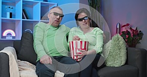 Older man and woman watching tv together, sitting at home on sofa watching a fascinating film in 3D glasses. Senior