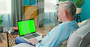 Older Man Works on a Laptop Computer with Green Mock-up Screen Sitting on a Couch