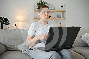 Older mature woman using wireless laptop apps browsing internet sit on sofa, smiling middle aged grandmother working