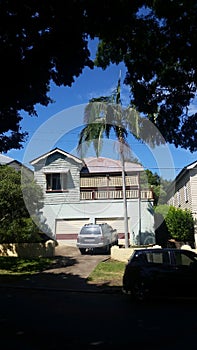 Older Queenslander house with cement block garage underneath and car parked in driveway framed by shade and greenery with a tall p