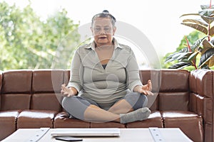 An older senior grey hair Asian woman sitting on sofa in living room and practicing basic yoga with calm and concentrate manner